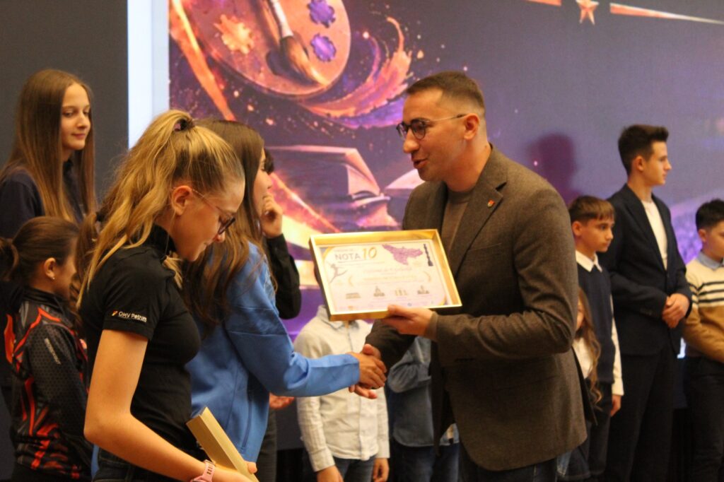 bărbat în sacou înmânează o diplomă „Nota 10” unei eleve, pe scenă
