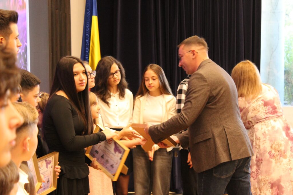 bărbat înmânează diplome și cutii unor eleve, pe fundal drapel românesc