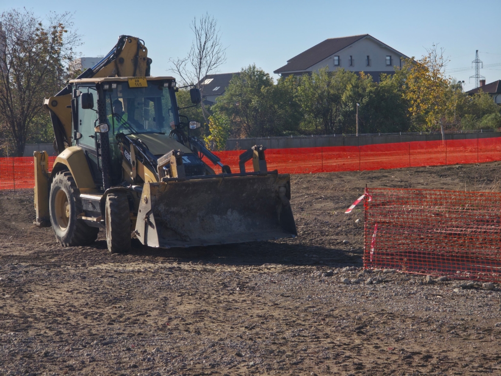 buldoexcavator pe un teren viran, delimitat cu plasă portocalie de șantier