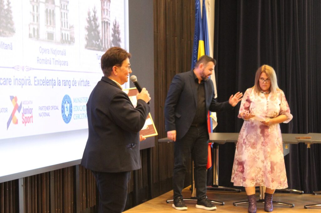 Trei persoane pe scenă, bărbat cu microfon și diplomă, lângă ecran de prezentare